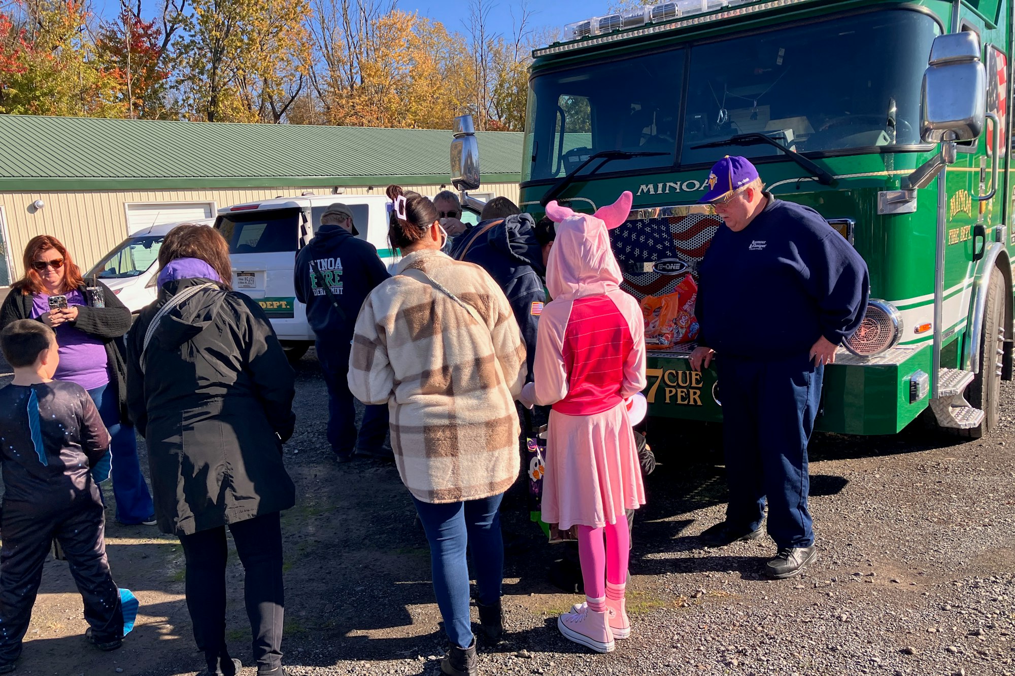Group of people gathered around a green firetruck on a sunny day, some in casual wear, one in a pink costume.