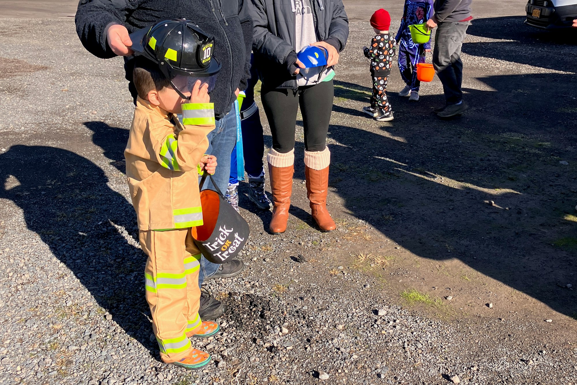 Child in firefighter costume with a helmet and trick-or-treat bucket, standing near adults on a sunny day.