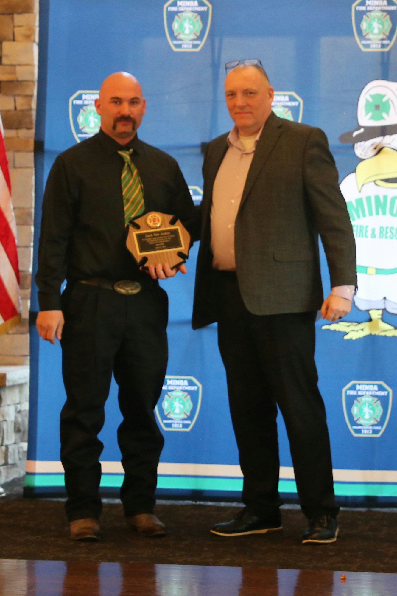 Two men are posing together; one is receiving an award. The backdrop features logos for a fire department event.