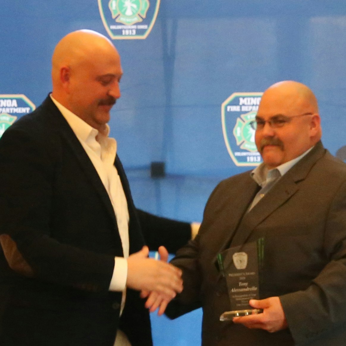 Two men are shaking hands at a ceremonial event, one presenting an award or plaque to the other. A logo background is visible.