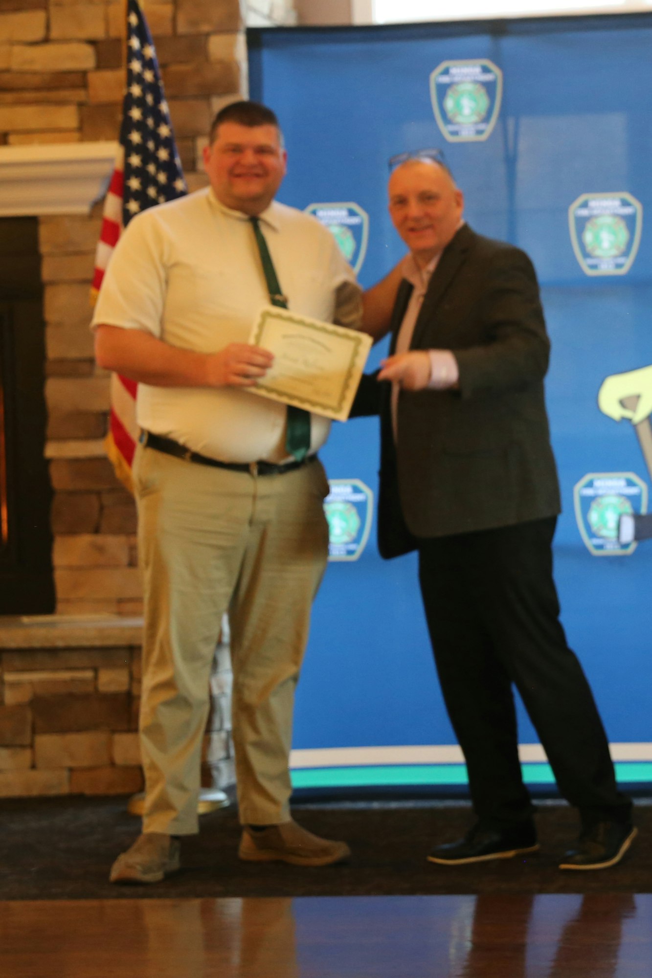 Two men are at a ceremony, one receiving a certificate while standing next to a banner and an American flag.