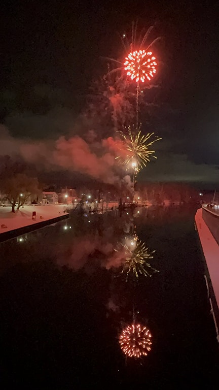 Fireworks illuminate the night sky, reflecting in a calm waterway, creating a festive and vibrant scene.
