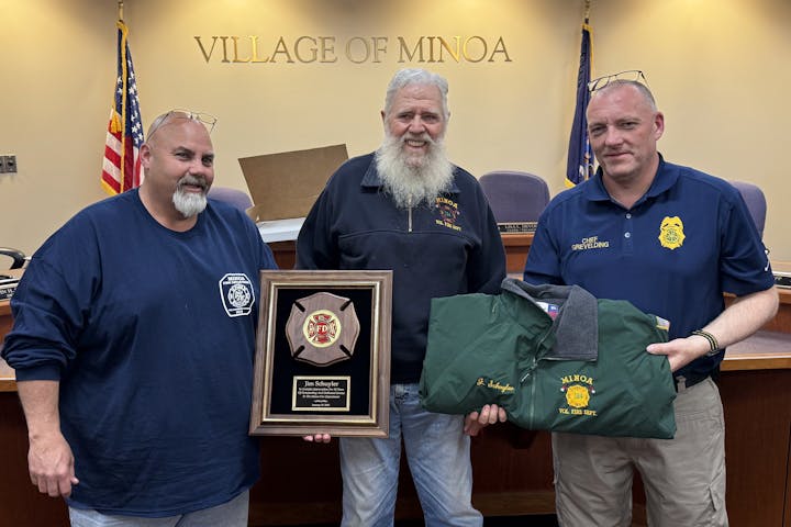 Tim Decker, Jim Schuyler, and Chief Grevelding pose with a plaque and a green jacket, under the sign "Village of Minoa."