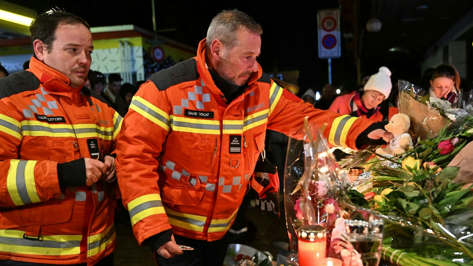 Firefighters in orange jackets are paying respects at a memorial adorned with flowers, candles, and a teddy bear.