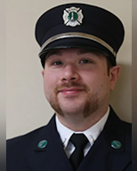 A person in uniform, wearing a cap with a badge, smiling at the camera. The setting appears to be indoors.