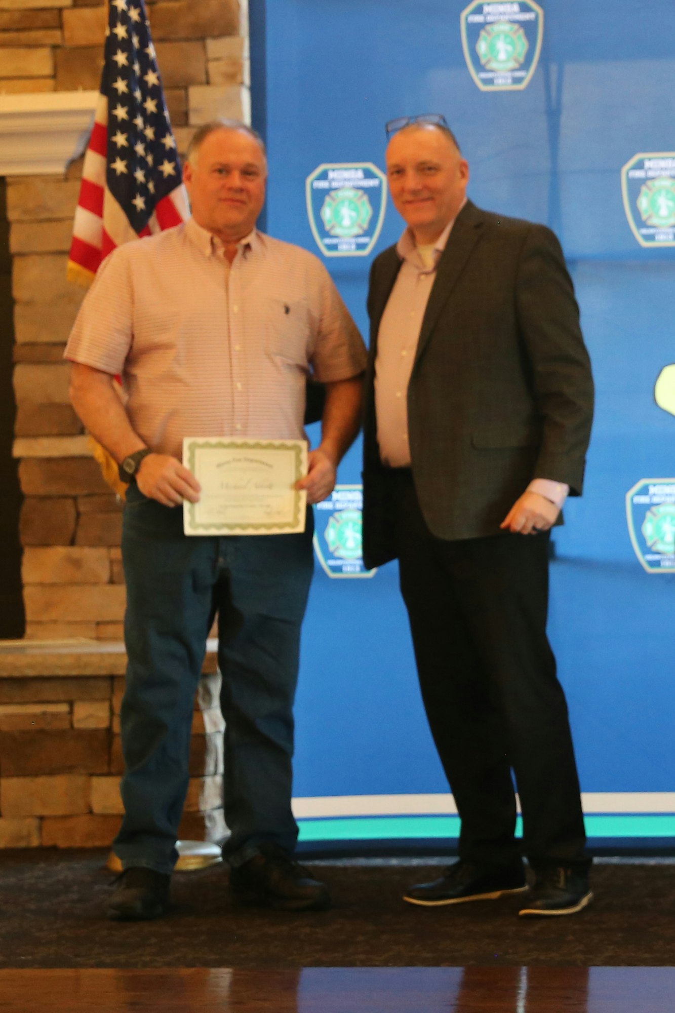 Two men stand together at an event, one holding a certificate, with an American flag and a blue backdrop in the background.