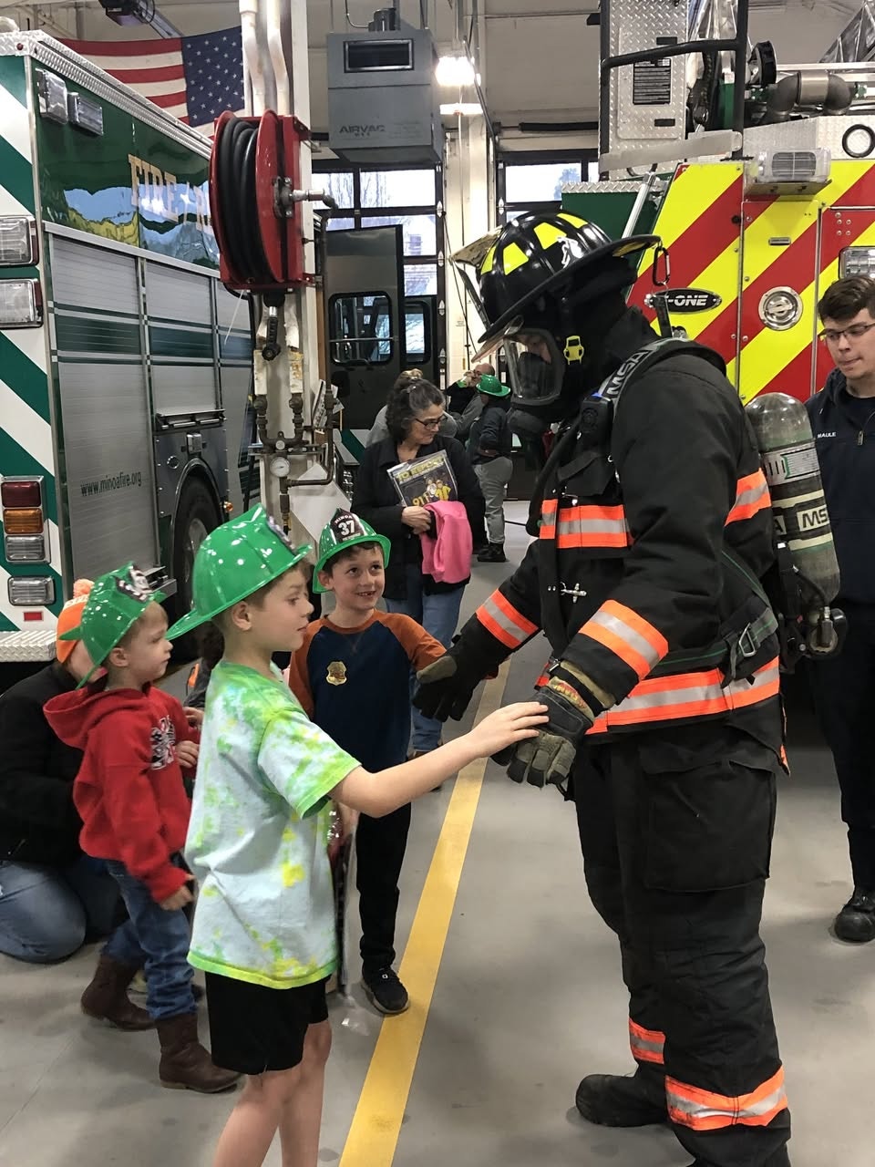 A firefighter interacts with children wearing green helmets in a fire station, with fire trucks visible in the background.