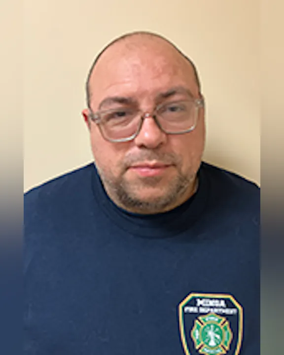 Tim Bennett wearing glasses and a dark shirt with a badge, standing against a plain background.