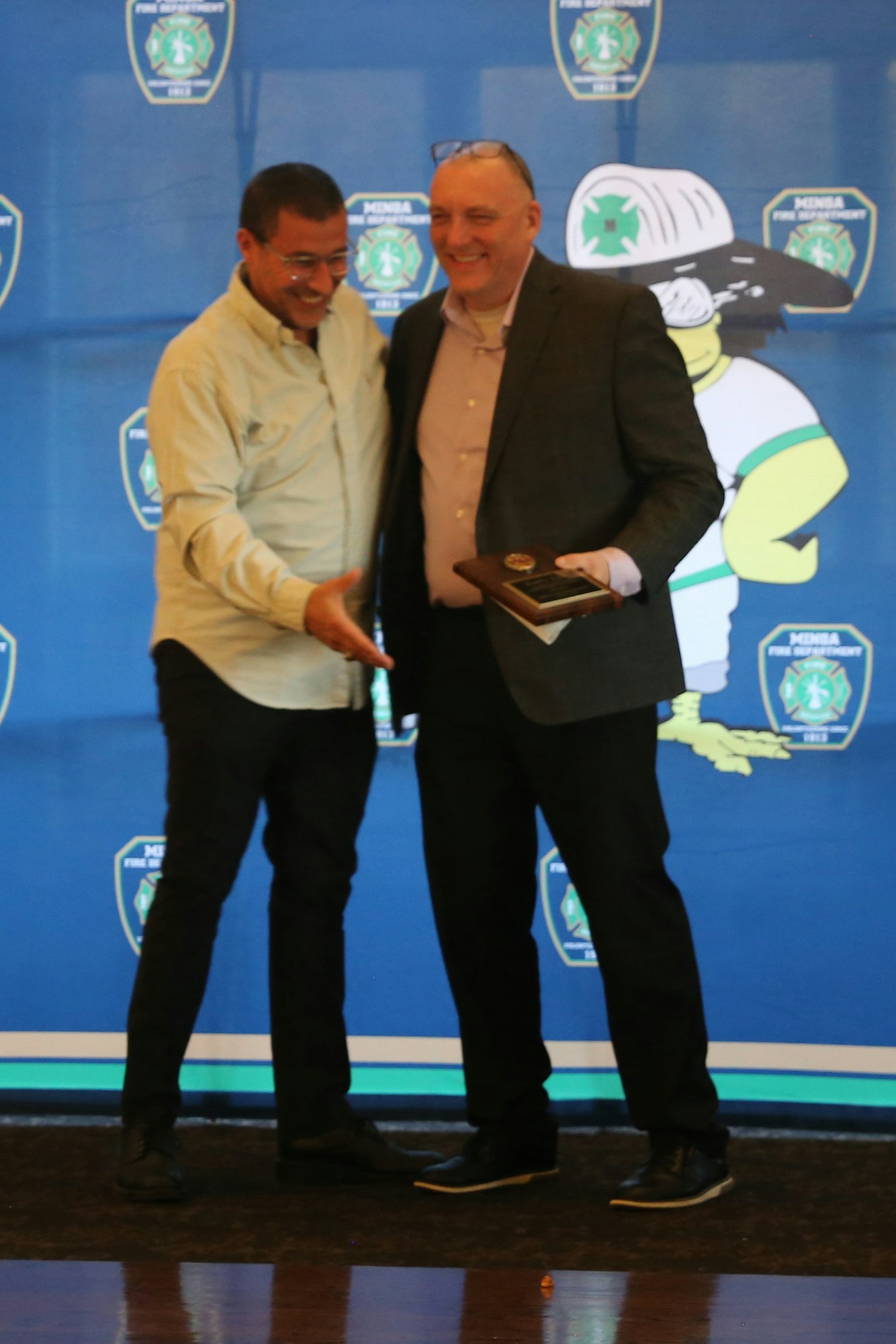 Two men are smiling and shaking hands at an event, with an awards plaque visible. A cartoonish firefighter character is in the background.
