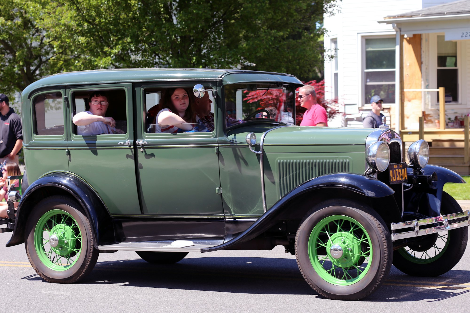 A vintage green car with green-spoked wheels driving on a street with people inside and others walking nearby.