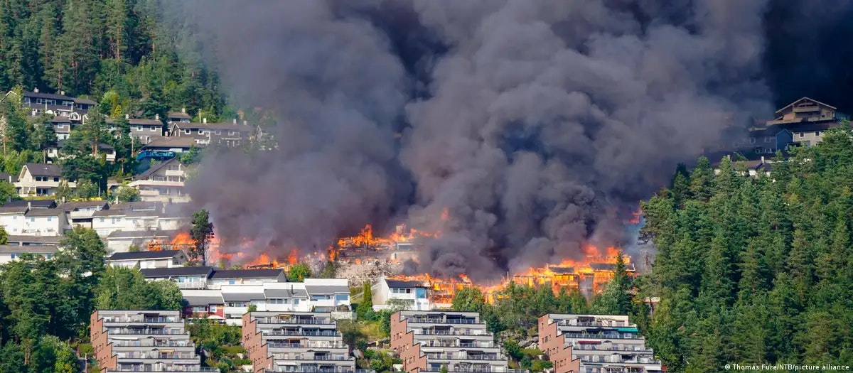 A large fire is consuming buildings, producing thick smoke, amidst a forested area and residential structures.