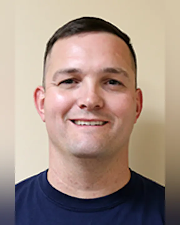 Tylor Cota with short hair, wearing a dark blue shirt against a beige background.