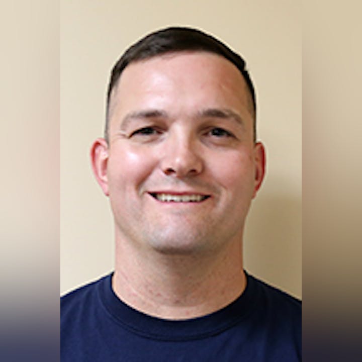 Tylor Cota with short hair, wearing a dark blue shirt against a beige background.