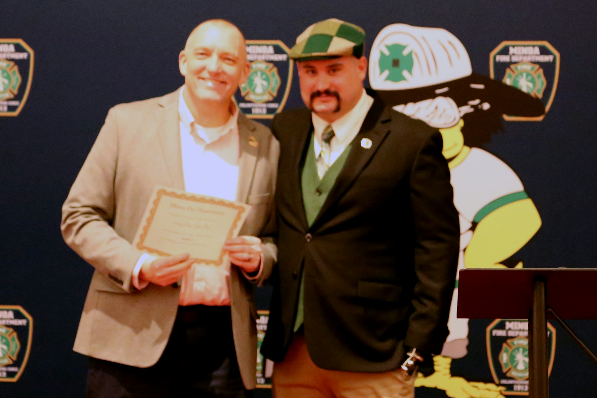Two people posing, one holds a certificate. Background has "Mina Fire Department" logos and a cartoon figure.