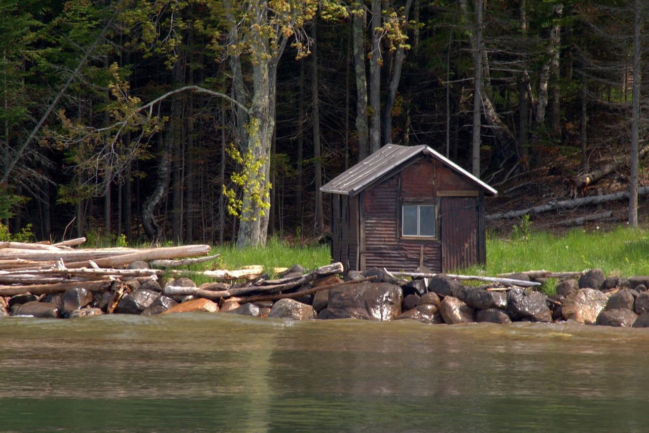 A small wooden cabin by a forested lakeside with rocks and driftwood.