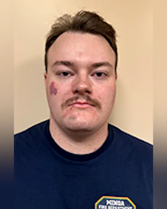 Zach Grevelding with short hair and a mustache, wearing a dark shirt with a badge-like symbol. Neutral background.