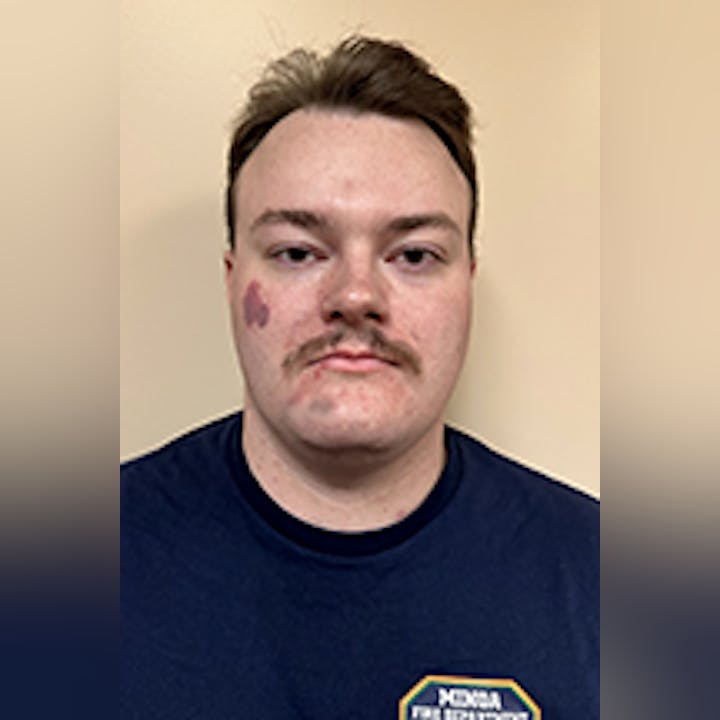 Zach Grevelding with short hair and a mustache, wearing a dark shirt with a badge-like symbol. Neutral background.