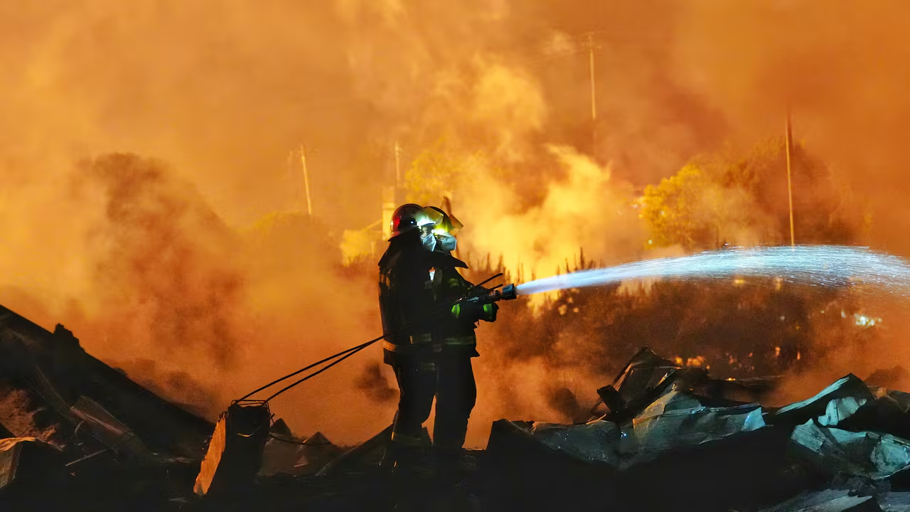 A firefighter battles a large blaze, spraying water amidst smoke and flames, highlighting the intensity of the firefighting effort.