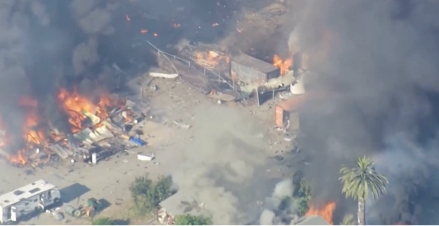Aerial view of a large fire with thick smoke, burning structures, debris, and a nearby palm tree.