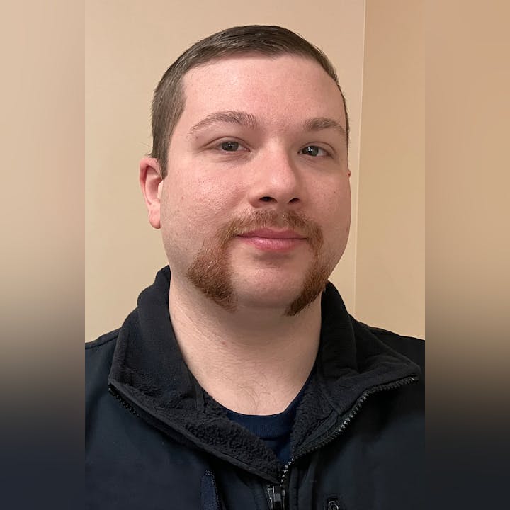 Vinny Caricofolo, with distinct mutton chops and a neutral expression, is wearing a dark jacket against a light background.