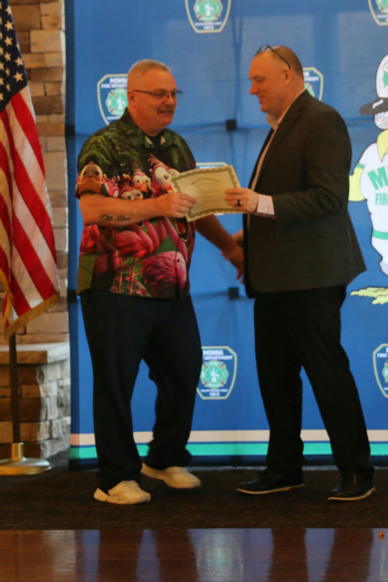 Two men are on stage, one in a colorful shirt receiving a certificate from the other, with an American flag in the background.