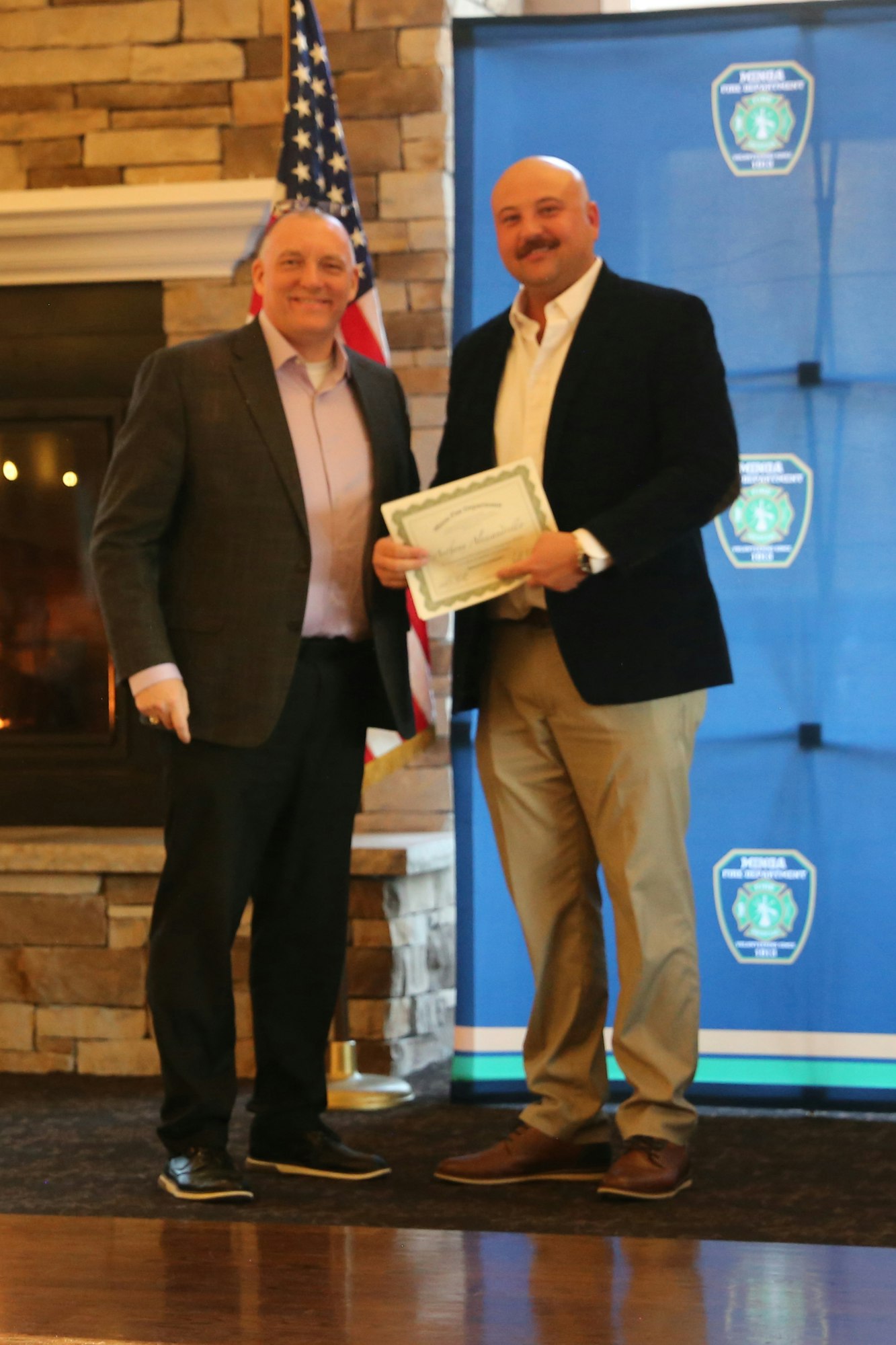 Two men stand together, one holding a certificate, in front of an American flag and a decorated backdrop, likely celebrating an achievement.