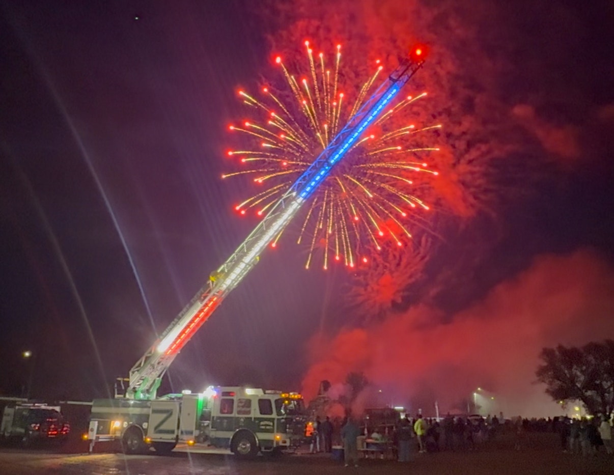 A Minoa fire truck with an extended ladder lit in red and blue, under a burst of red fireworks in a nighttime scene with a crowd below.