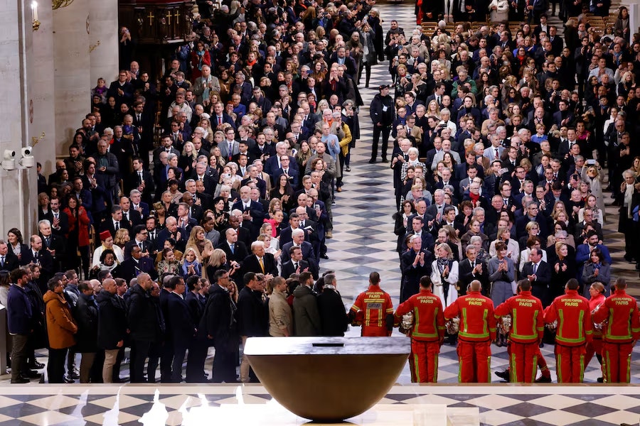 A large crowd of people in formal attire gathered for the reopening of Notre Dame Cathedral, with firefighters in uniform standing at the front.
