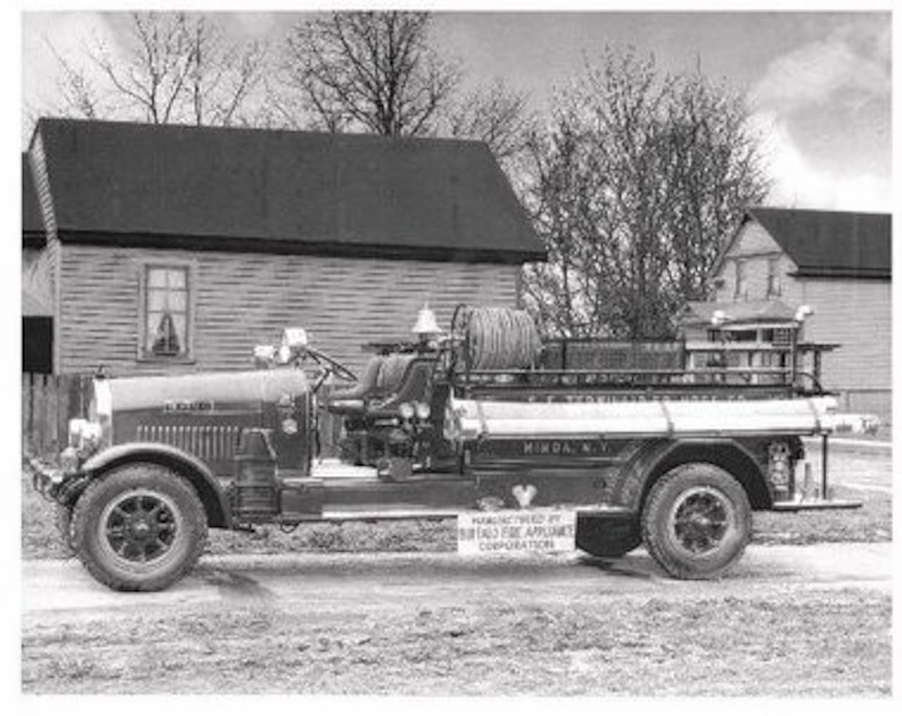 A vintage fire truck parked in front of old wooden houses, with trees in the background.