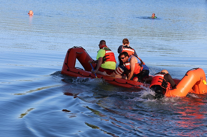 People in life vests are on an inflatable raft, with some in the water nearby.