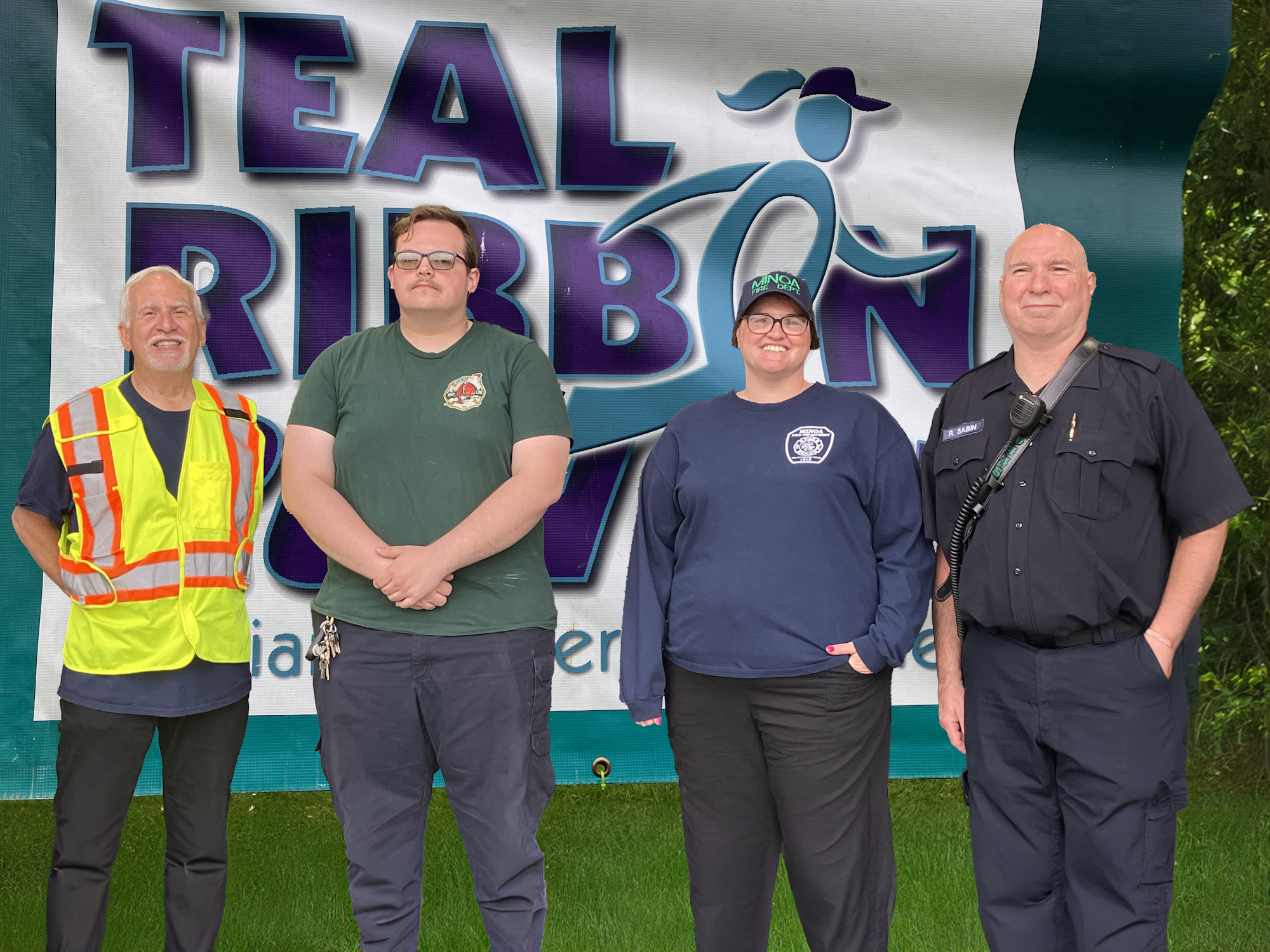 Four volunteers of the Minoa Fire Department posing in front of a "TEAL RIBBON" sign, representing different professions; one in a safety vest, another in casual attire, the third and fourth in uniform.