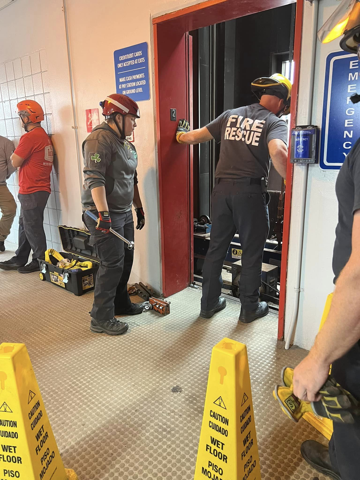 Firefighters with helmets and tools inside a building with "CAUTION WET FLOOR" signs.