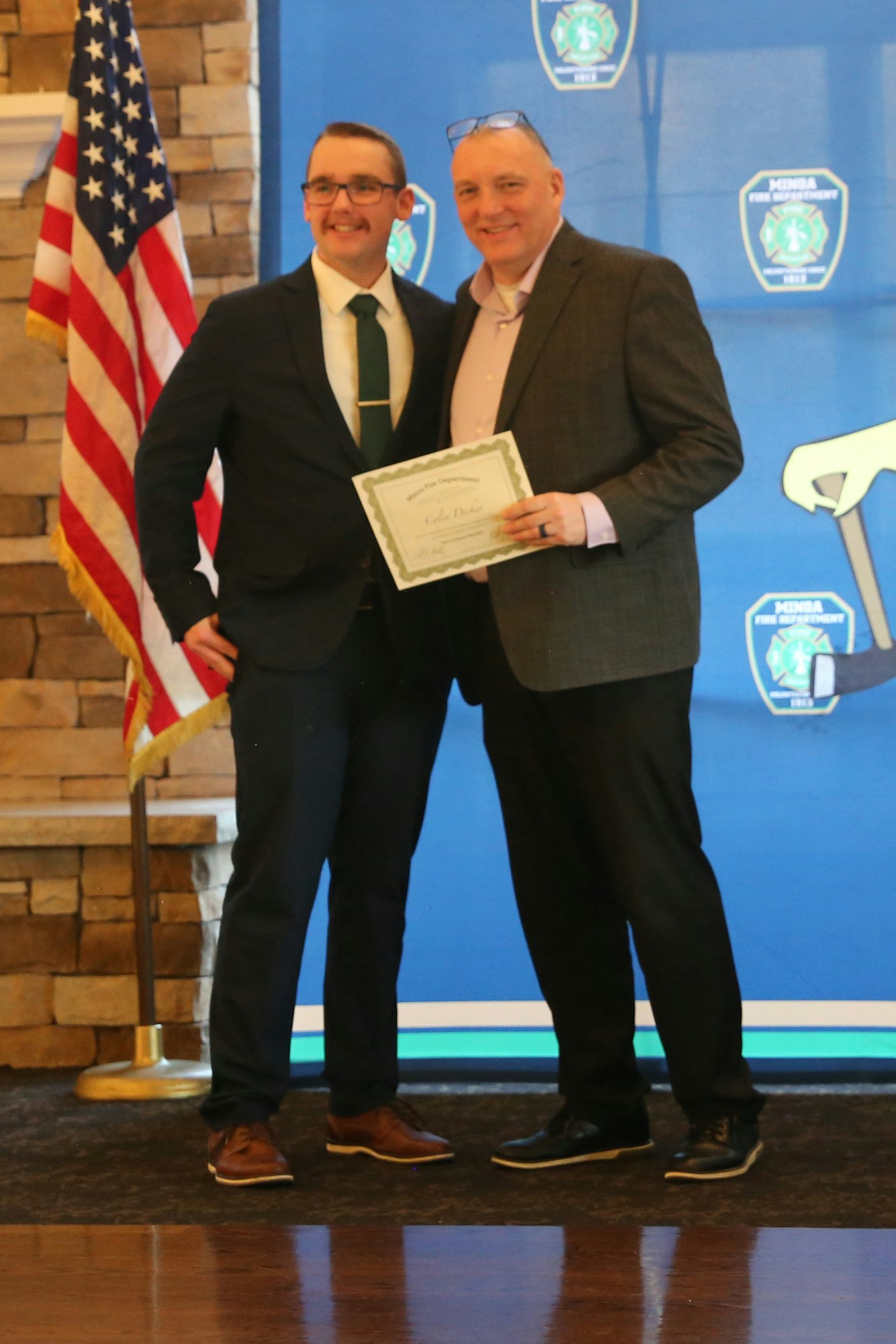 Two men are smiling for a photo, one holding a certificate, with an American flag in the background.