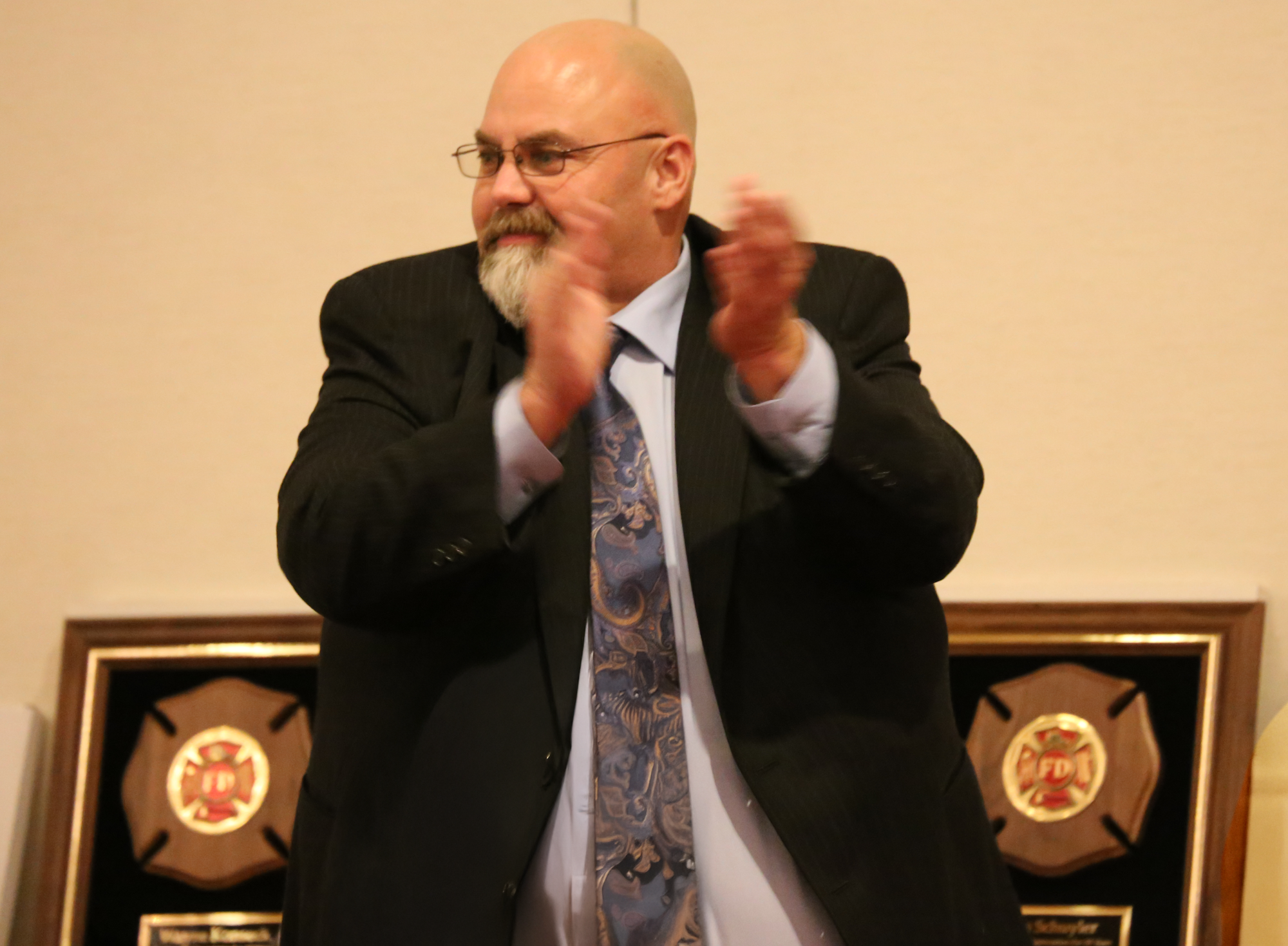 President Tim Decker in a suit clapping with fire department plaques in the background.