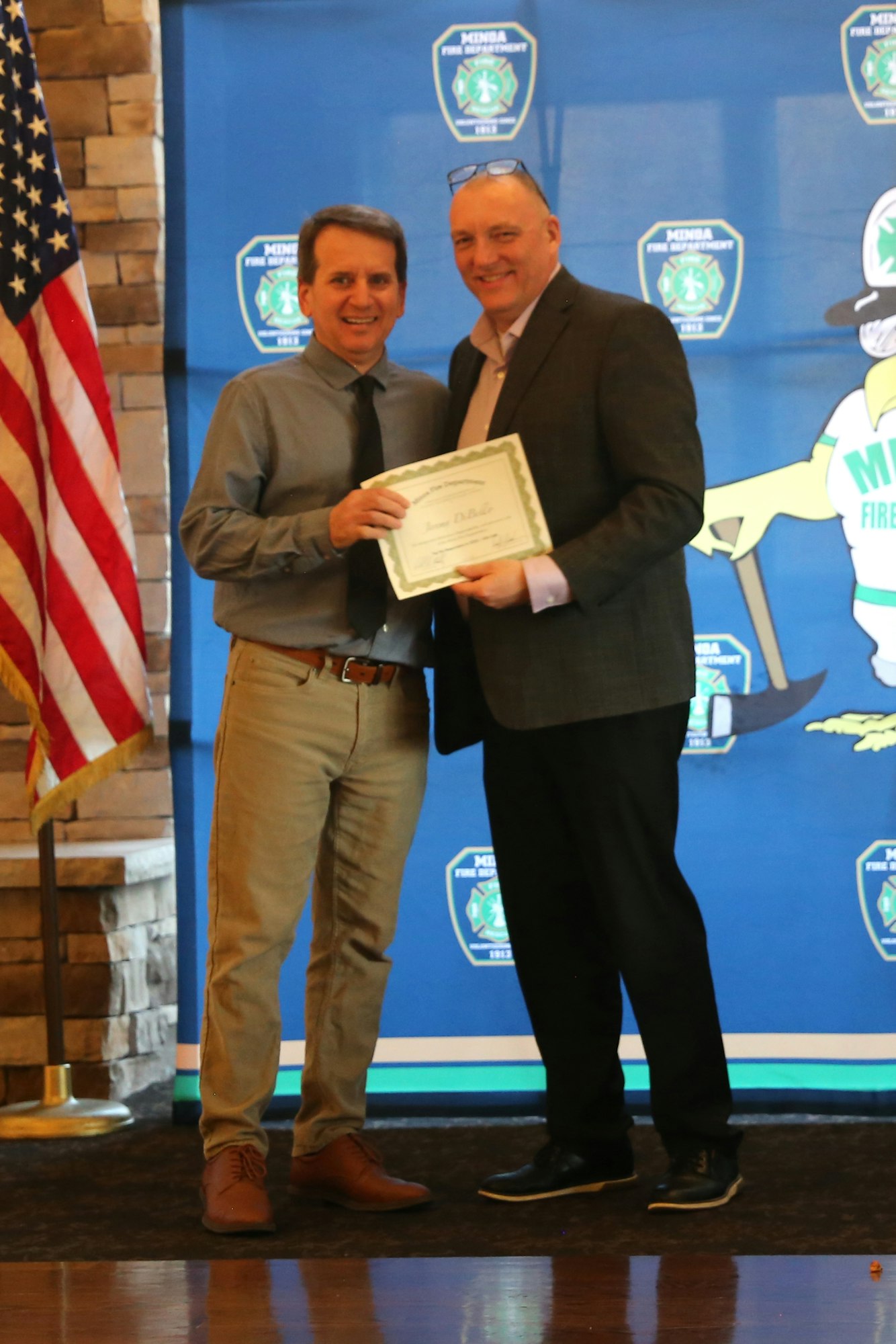Two men are smiling and shaking hands while holding a certificate, with an American flag and a fire department backdrop.