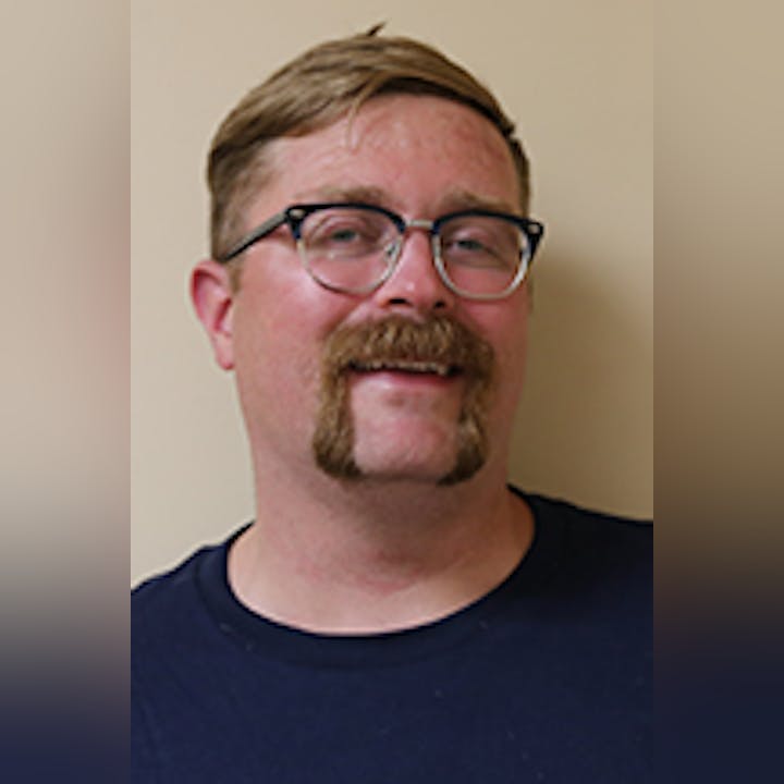 Stephen Phillis with glasses and a mustache, smiling against a plain background.