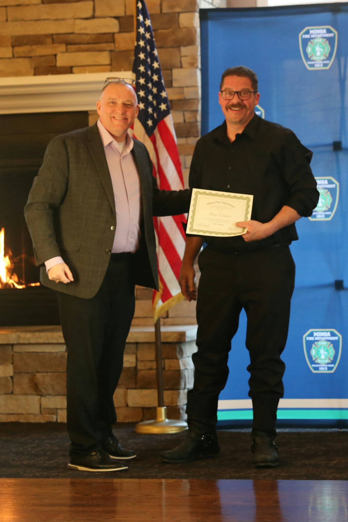 Two men are smiling and posing for a photo, one holding a certificate, with an American flag and decoration in the background.