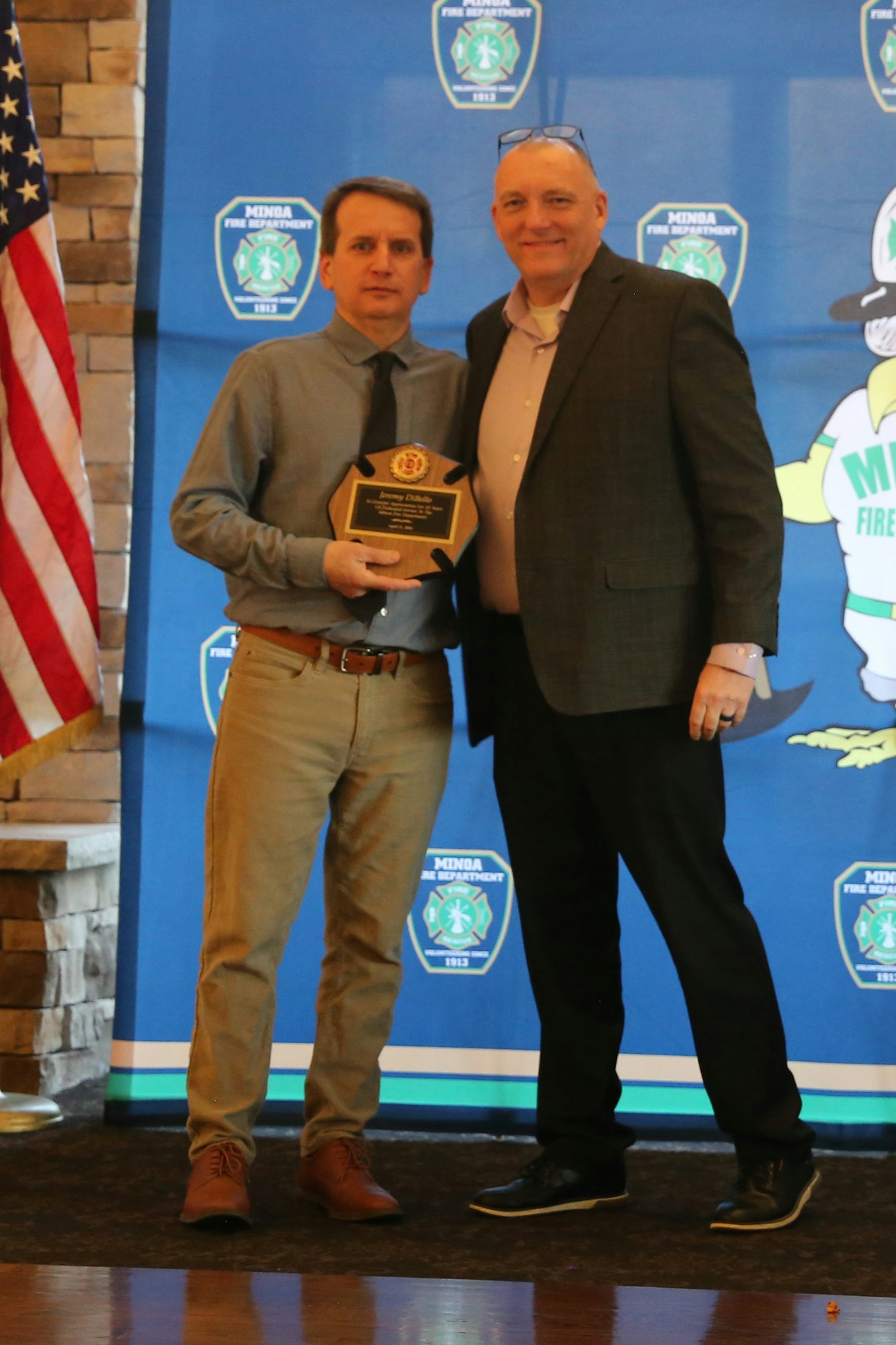 Two men are posing together, one holding an award plaque, with a banner for a fire department in the background.