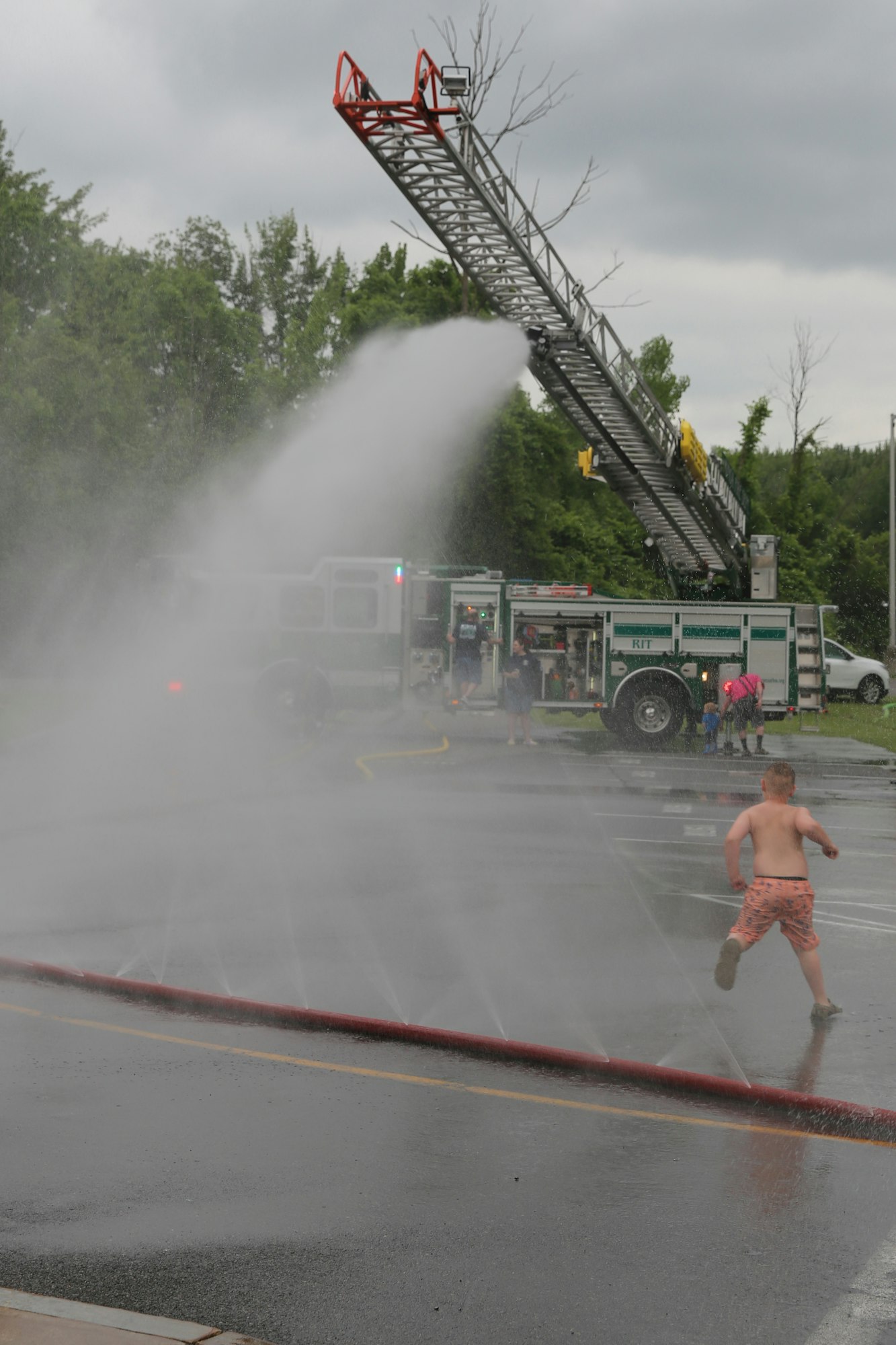 Fire truck spraying water; a shirtless child runs through the spray.