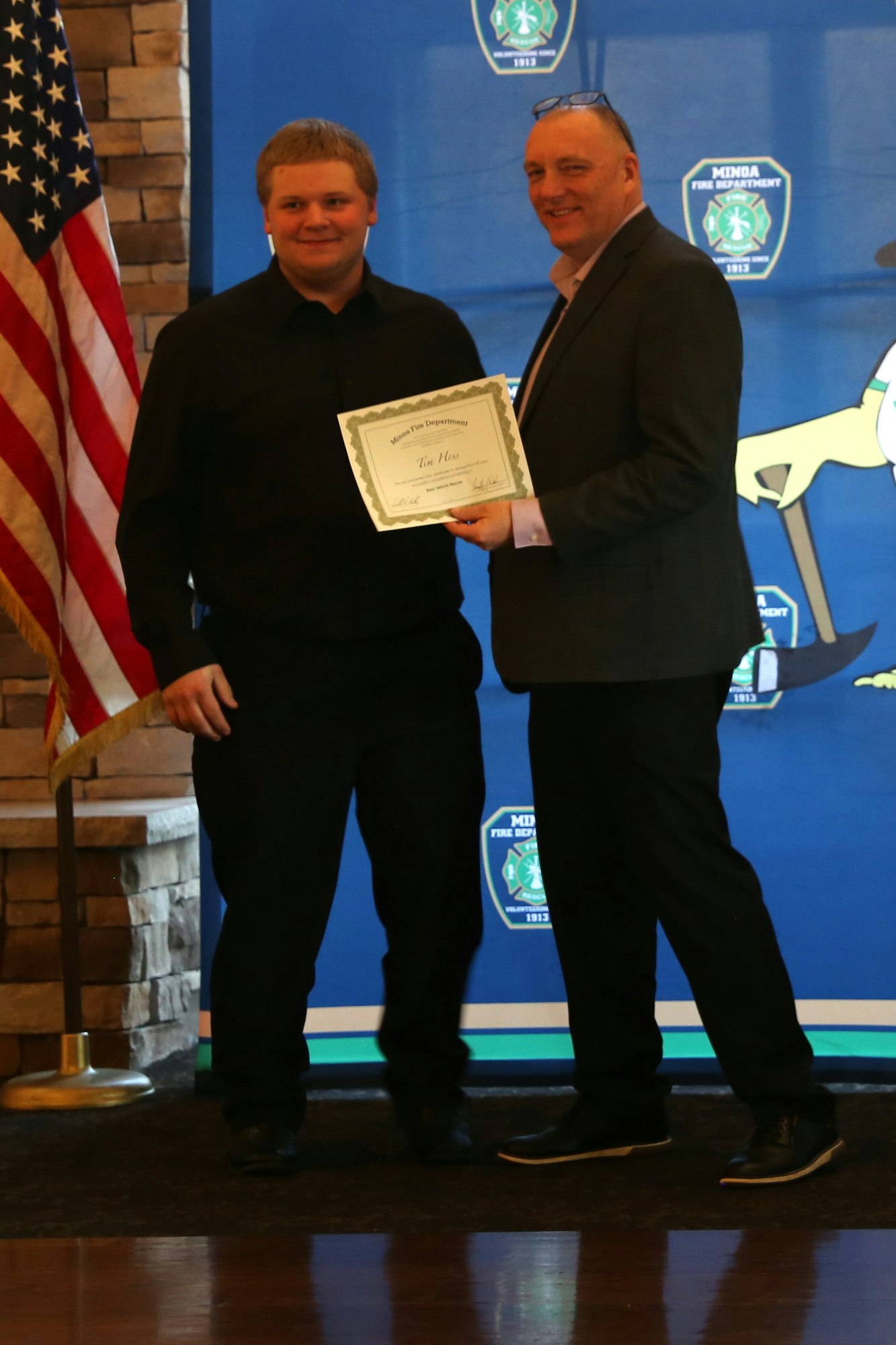 Two men stand together, one holding a certificate, with an American flag in the background and a themed backdrop behind them.