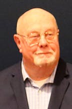 Jack Robie with glasses, wearing a suit jacket and a collared shirt, standing against a dark background.