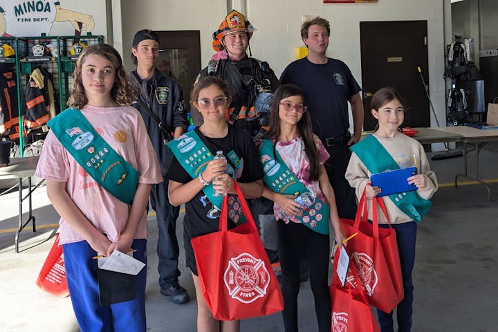 A group of Girl Scouts with red "Prevent Fires" bags posing with firefighters at a fire station.