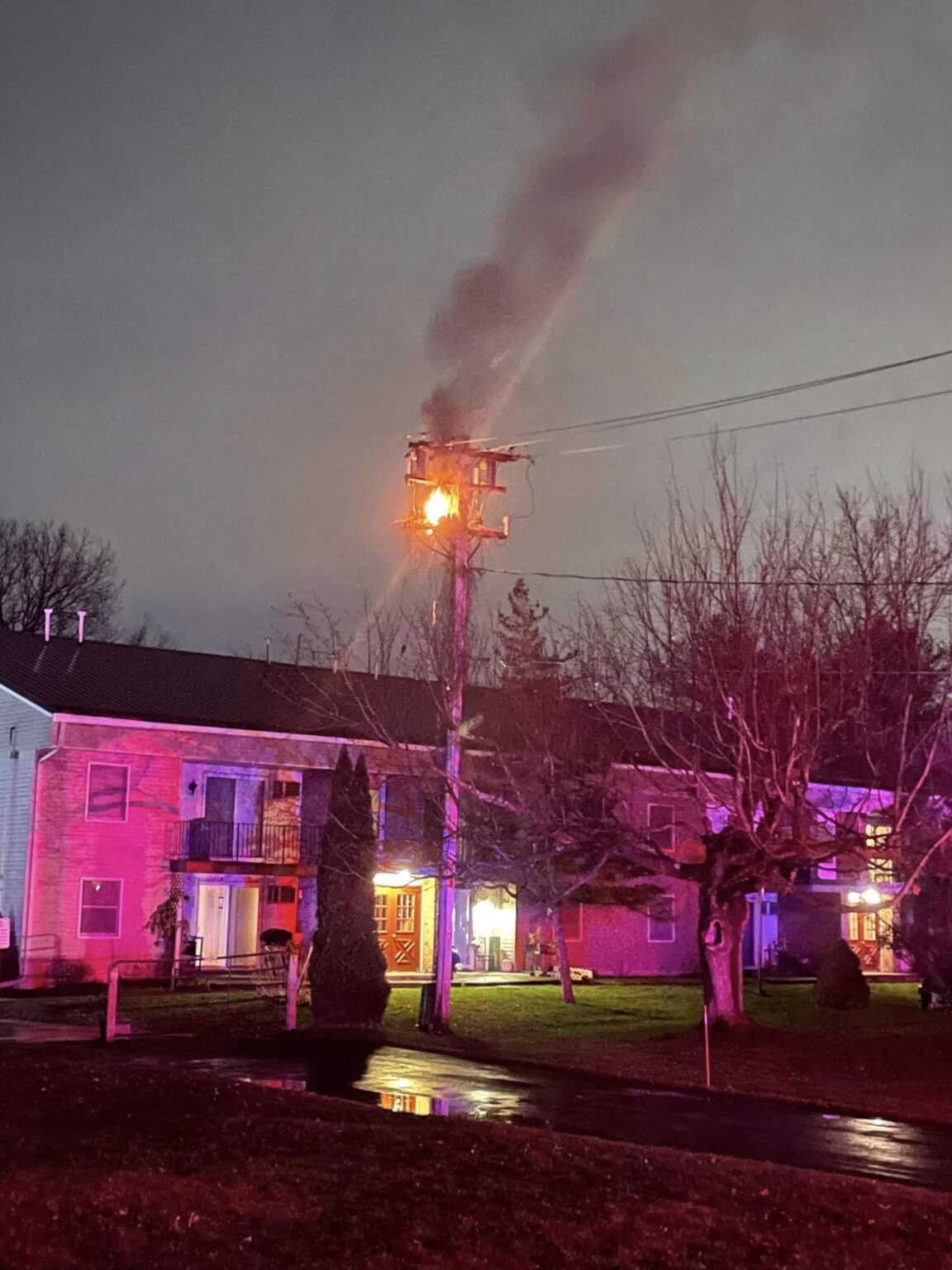 A utility pole on fire at night with smoke, near residential buildings.