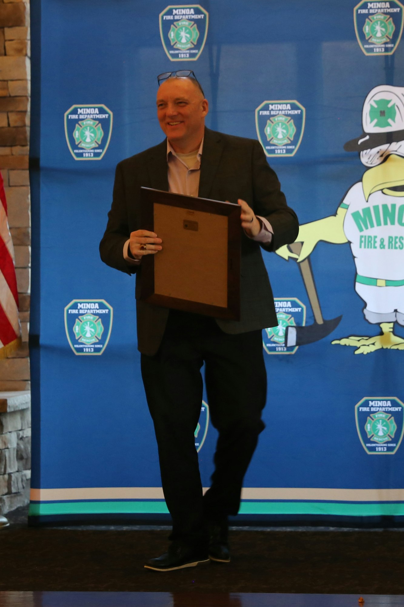 A smiling man holds a frame, walking in front of a Minda Fire Department backdrop at an event, with an American flag nearby.