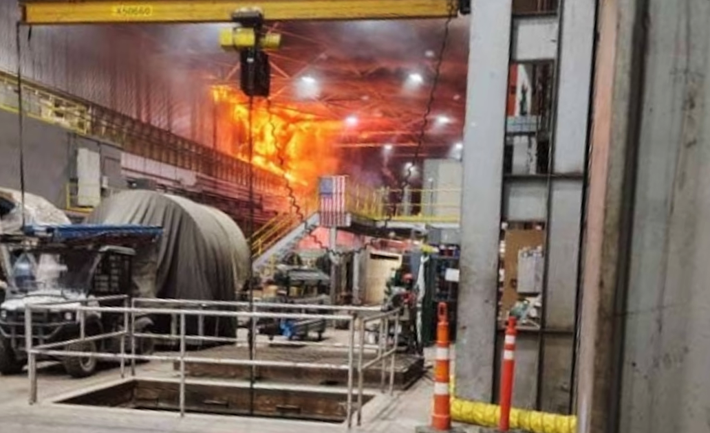 A large fire inside the Novelis industrial facility, with visible flames and smoke, surrounded by equipment and safety barriers.