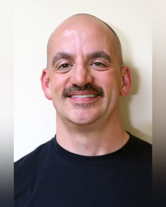 Luke Bellandi, a smiling bald man with a mustache, wearing a black shirt, against a light-colored background.