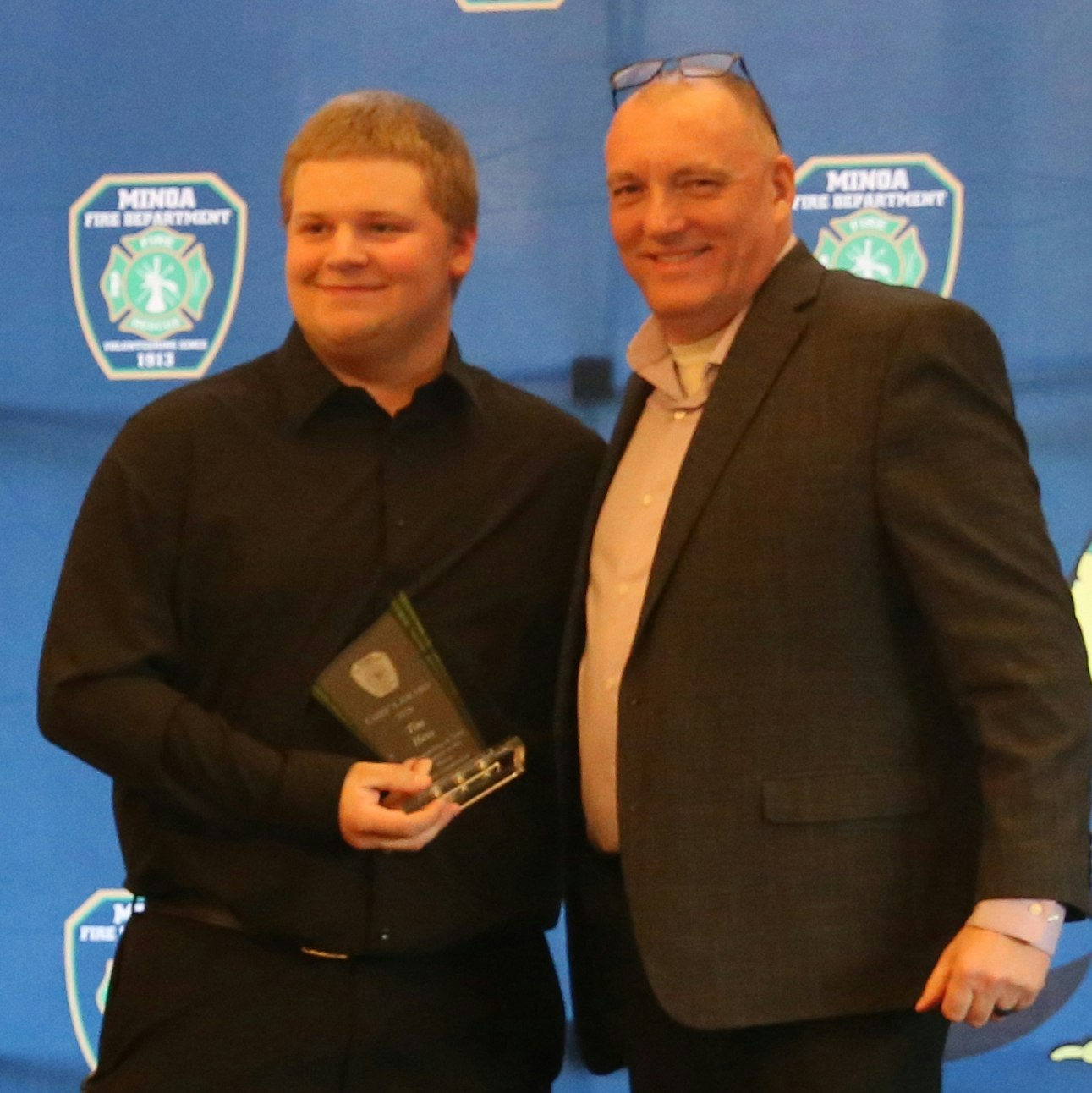 Two men pose for a photo, one holding an award. The backdrop features the logo of the Minda Fire Department.
