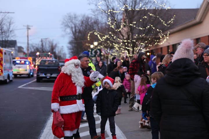 Santa and a crowd at a festive street event, surrounded by decorated trees and emergency vehicles.