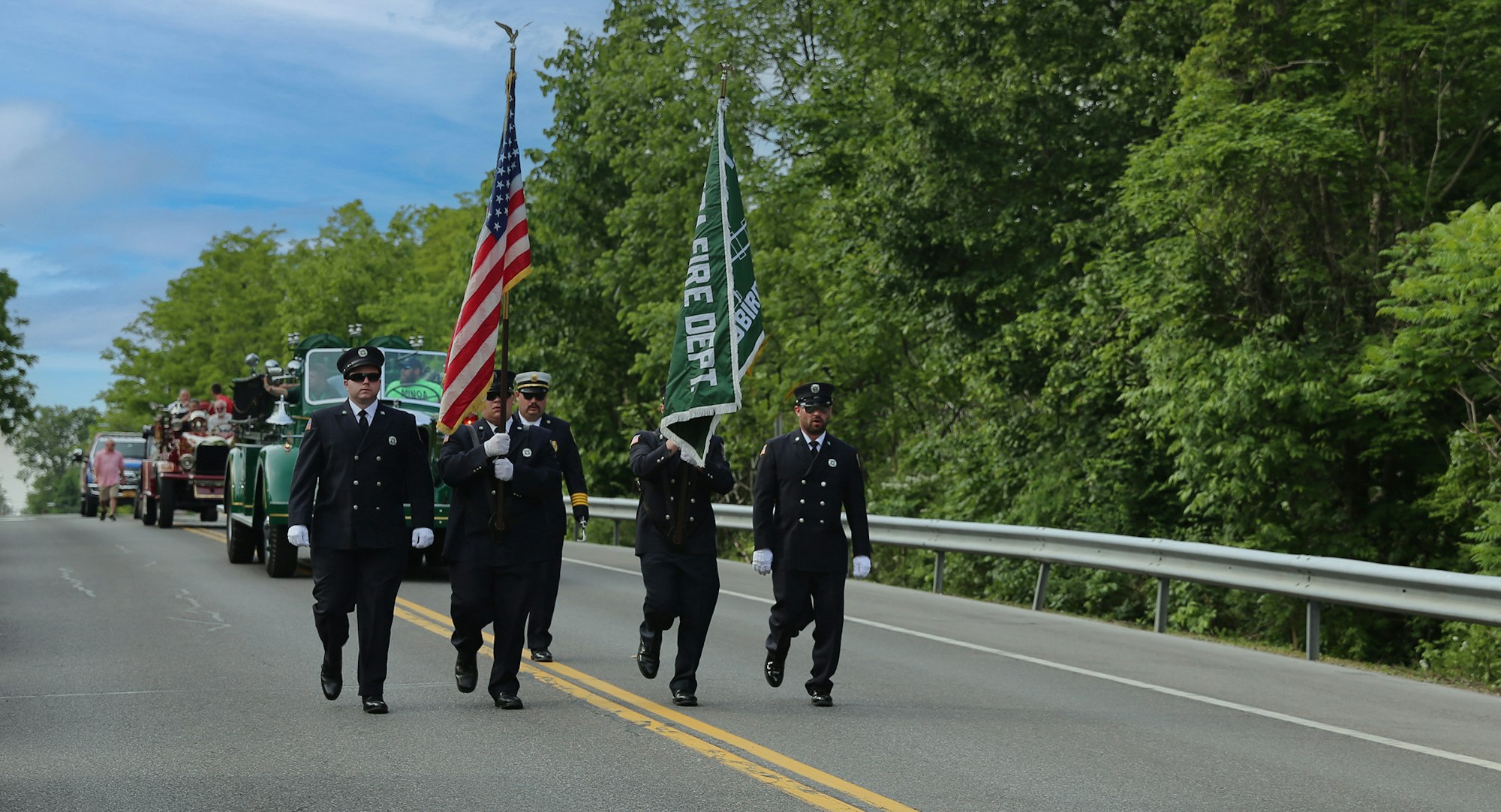 A procession of Minoa firefighters carrying flags lead a parred along a tree-lined road.