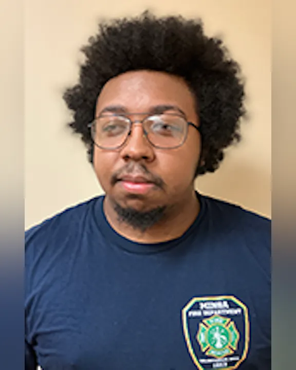 Zachariah Sewell with curly hair and glasses, wearing a T-shirt with a patch on it.